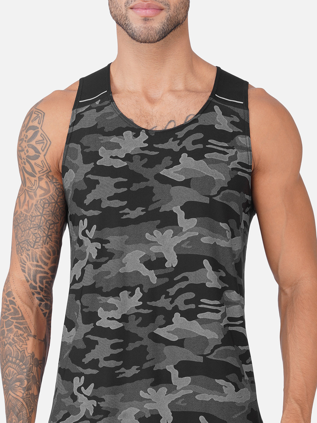 A man wearing a sleeveless tank top with a black and gray camouflage pattern, shown from the chest down against a light gray background.