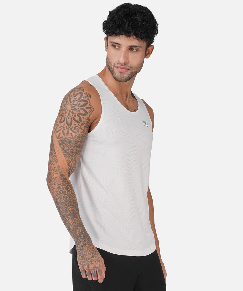 A man with curly black hair and a tattooed left arm wearing a white ribbed sleeveless tank top and black pants, photographed against a light gray studio background.