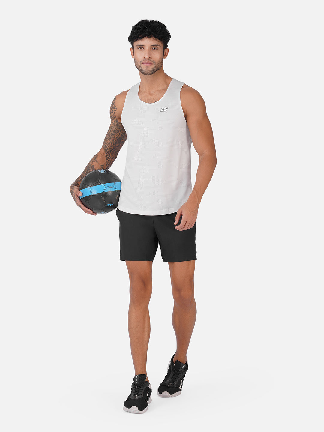 A man modeling a white athletic tank top and black shorts, holding a black and blue medicine ball against a light gray studio background.