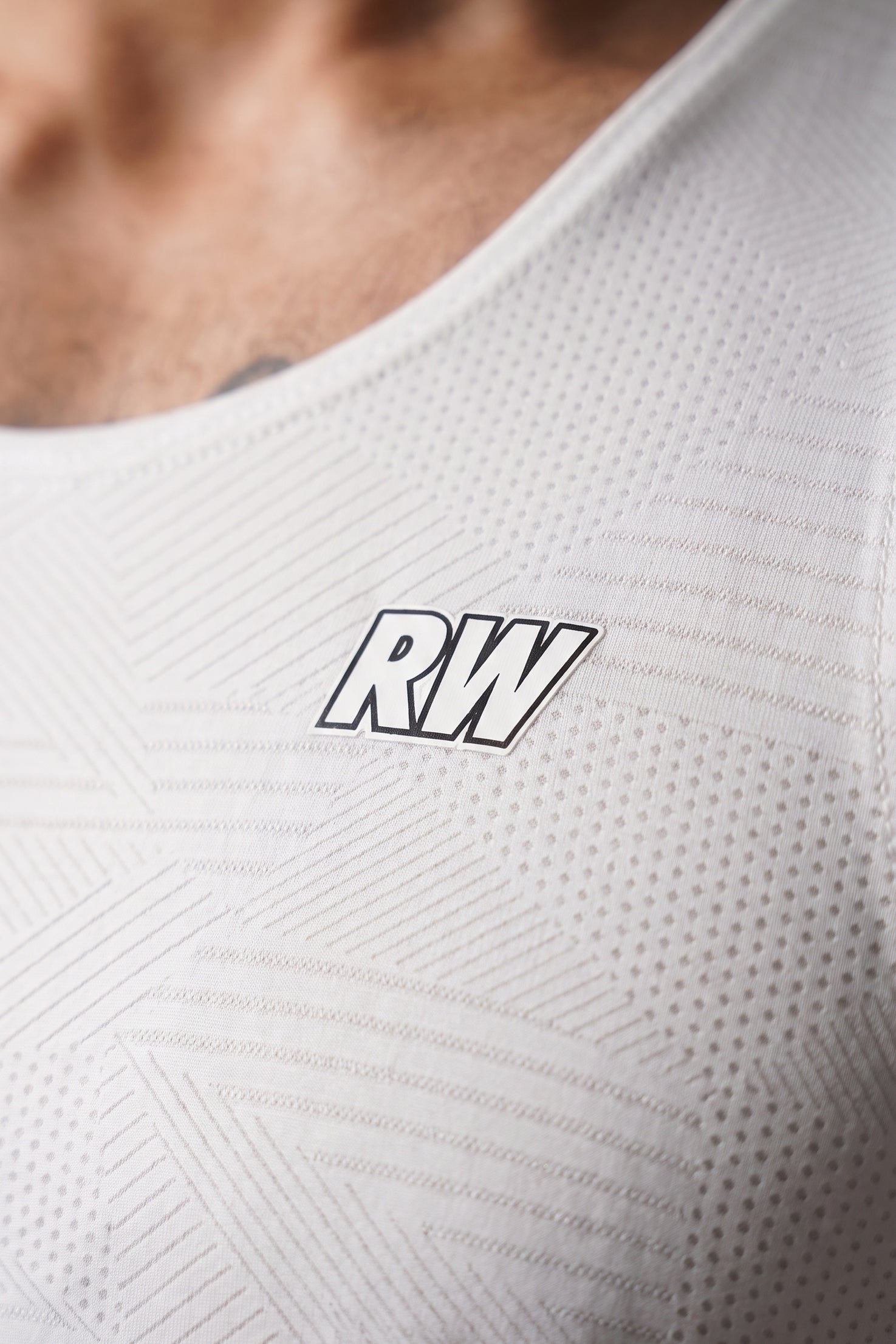 Close-up of a white athletic tank top with a textured geometric pattern and a black outlined 'RW' logo on the chest, worn by a model.