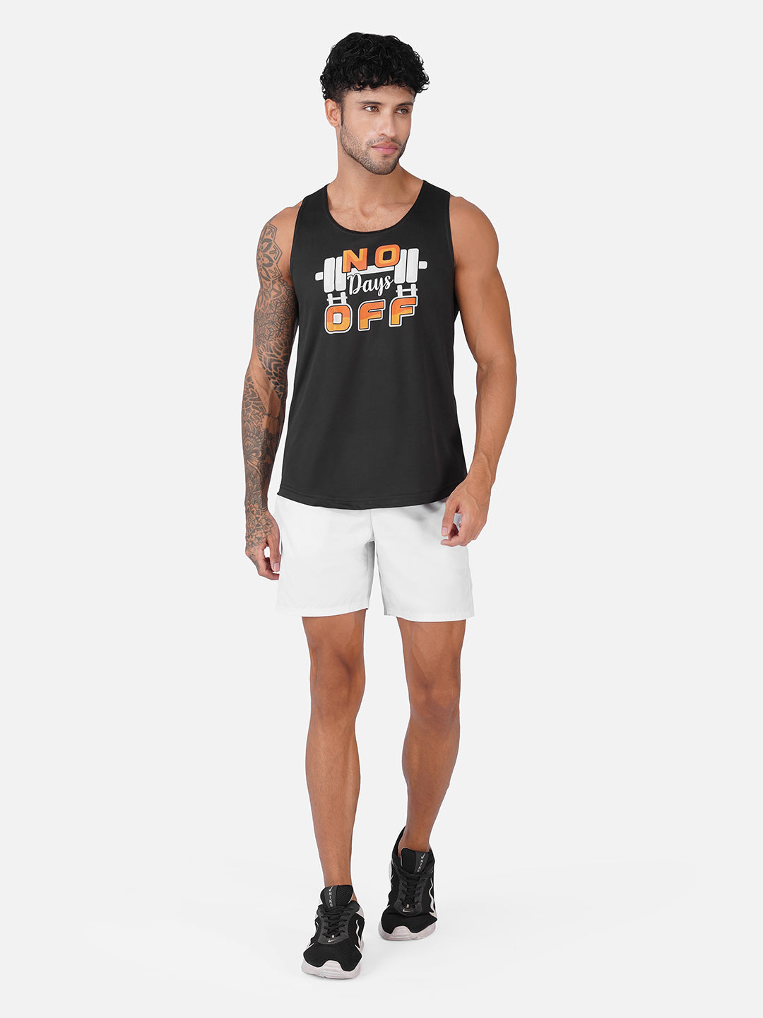 A man modeling a black sleeveless tank top with a white and orange graphic that reads 'No Days Off' with barbell details, paired with white shorts and black sneakers, against a light gray studio background.