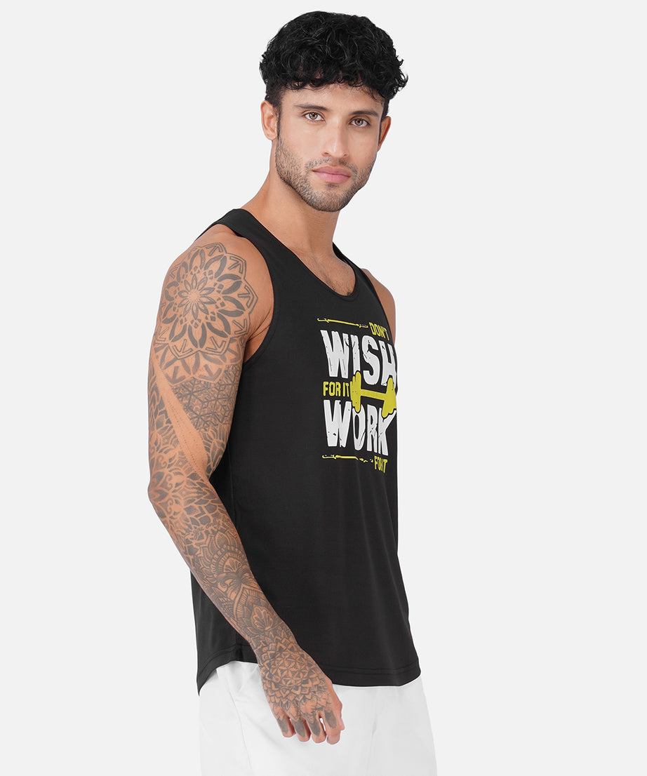 A man wearing a black sleeveless tank top with a yellow and white graphic print that reads 'WISH FOR IT WORK', paired with white pants, photographed against a light gray studio background.