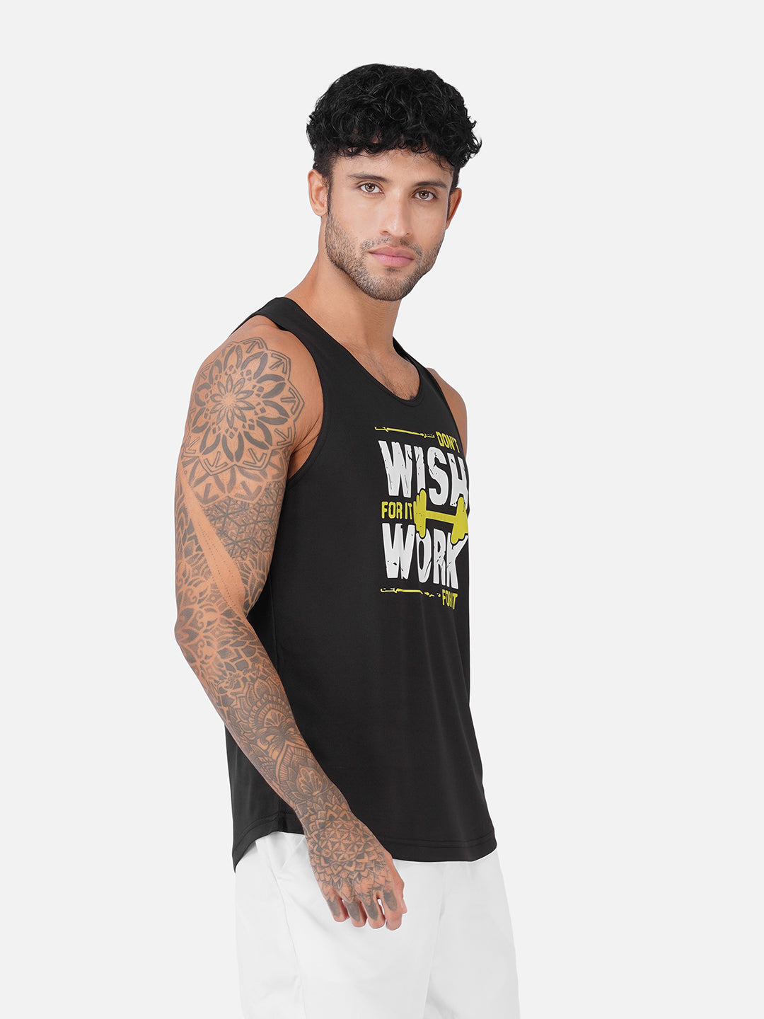 A man wearing a black sleeveless tank top with a yellow and white graphic print that reads 'WISH FOR IT WORK', paired with white pants, photographed against a light gray studio background.